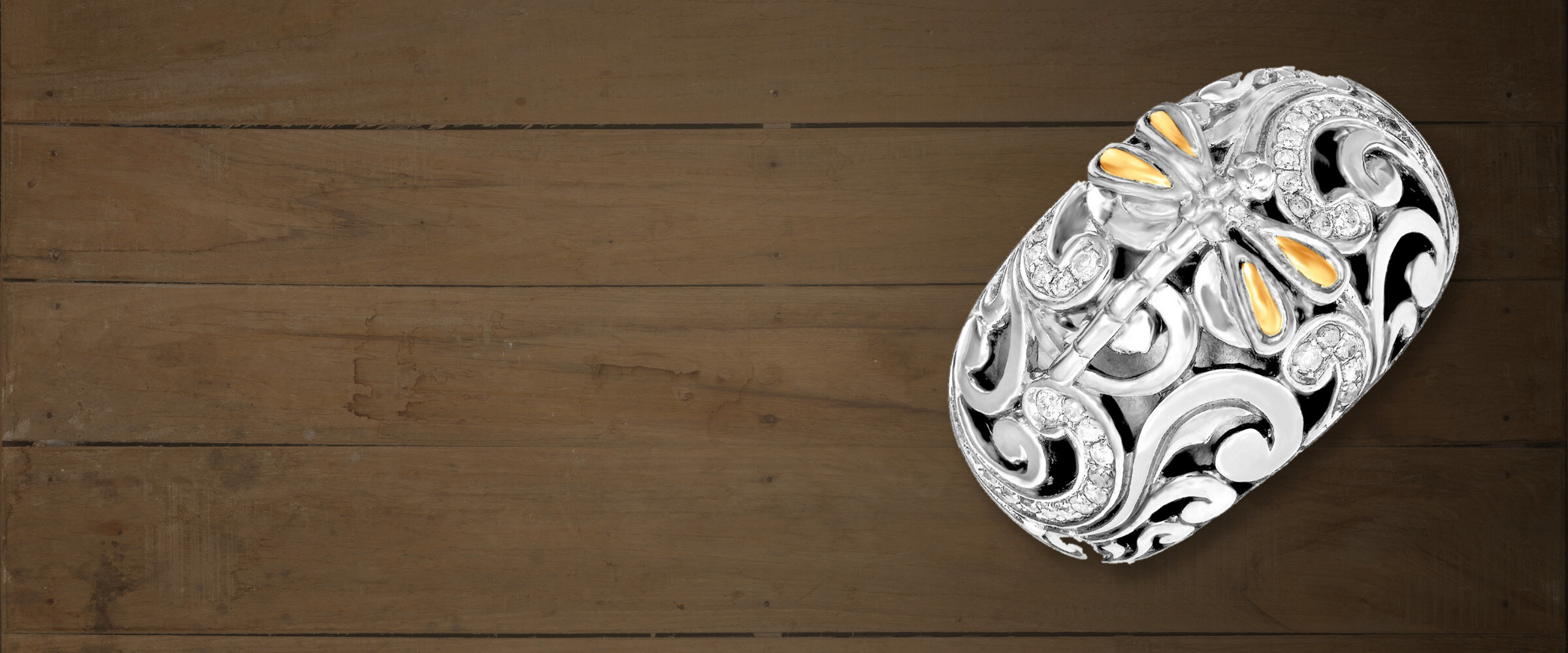 A sterling silver dome ring with an 18k gold dragonfly, filigree scrollwork and pave diamonds, resting on weathered wood planks